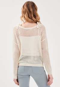 Pull léger en tricot blanc crémeux avec une trame lâche et ouverte. Présente un large décolleté et des détails de bretelles à nouer. Texturé et décontracté.