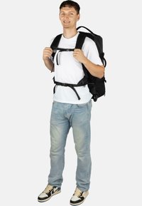 Schwarzer Rucksack mit verstellbaren Riemen, verschiedenen sicheren Schnallen und einem schlanken Design, getragen von einer Person in einem weißen Hemd und Jeans.