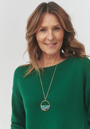 Green knitted sweater with ribbed neckline, featuring a circular gold pendant necklace displaying blue and gold abstract patterns.