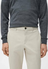 Mężczyzna w beżowych spodniach na guziki i z zamkiem błyskawicznym oraz w ciemnoszarym swetrze z długim rękawem, stojący z rękami swobodnie opadającymi wzdłuż ciała.