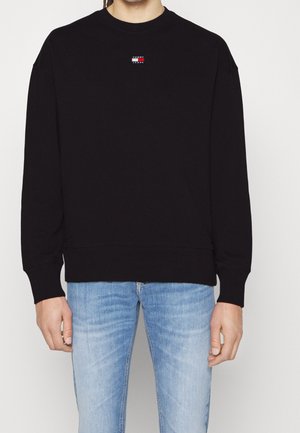 Person trägt einen schwarzen Langarmpullover mit kleinem Logo auf der Brust und hellblaue Jeans, steht vor einem einfarbigen Hintergrund.