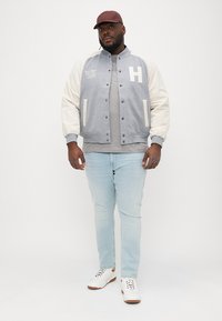 Jachetă varsity gri și crem cu butoane snap, având un mare "H" pe piept, asortată cu blugi albaștri deschiși și adidași albi.