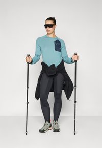 Hellblaues Langarmhemd mit dunkler Tasche, schwarze Leggings, graue Wanderschuhe und Sonnenbrille. Hält schwarze Trekkingstöcke.