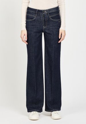 Frau trägt dunkelblaue, hochtaillierte, weite Jeans mit vorderen Knöpfen, ein weißes Langarmoberteil und weiße Sneakers und steht vor einem weißen Hintergrund.
