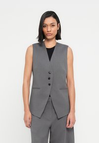 Γκρι αμάνικο γιλέκο από υφασμάτινη υφή, με βαθύ V λαιμόκοψη, τρία μαύρα κουμπιά και πλαϊνές τσέπες. Συνδυάζεται με γκρι παντελόνια.