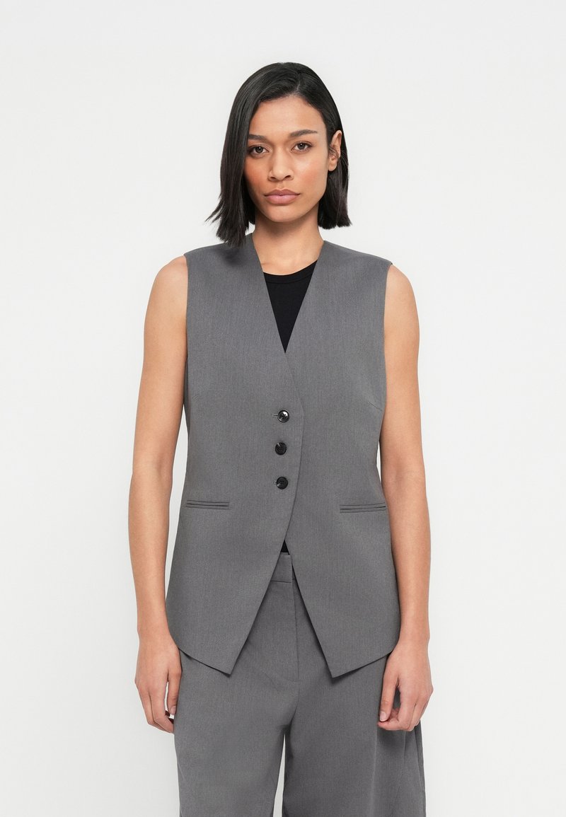 Γκρι αμάνικο γιλέκο από υφασμάτινη υφή, με βαθύ V λαιμόκοψη, τρία μαύρα κουμπιά και πλαϊνές τσέπες. Συνδυάζεται με γκρι παντελόνια.