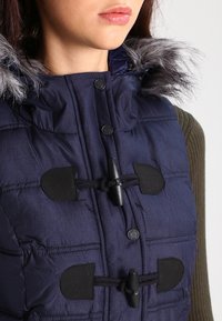 Marinblå vadderad väst med huva i fuskpäls, har stora knappar och snörningsknäppningar, samt ett strukturerat tyg med quiltade sektioner.