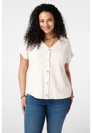 Vrouw met krullend haar, gekleed in een wit, kort mouwtjes overhemd en blauwe jeans, glimlachend tegen een effen achtergrond.