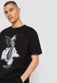 Svart bomull T-skjorte med et grafisk motiv av en vinget figur på en sky, med hvit tekst under designet. Standard rund hals og korte ermer.