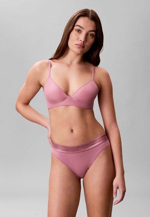 Femme aux longs cheveux bruns portant un soutien-gorge Calvin Klein mauve assorti et une culotte taille haute sur un fond gris uni.