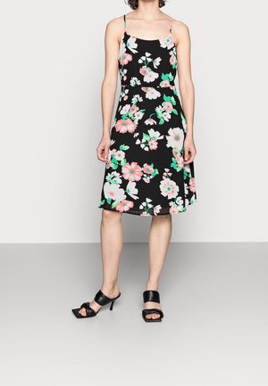 Kvinde iført sort ærmeløs knælængde kjole med pink og hvidt blomsterprint og sorte åbne hælede sandaler.