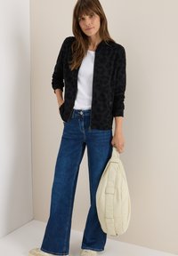 Veste bomber à imprimé léopard noir et gris, t-shirt blanc et jean évasé bleu. Tenant un sac à épaule rembourré de couleur crème.