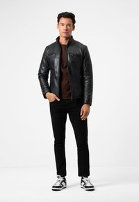 Schwarze Lederjacke mit Stehkragen, zwei Reißverschluss-Taschen, kombiniert mit einem braunen Hemd und schwarzen Jeans, getragen mit schwarz-weißen Sneakers.