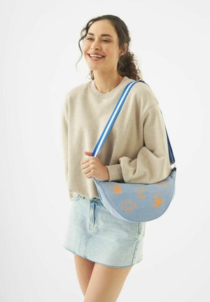 Junge Frau lächelt, trägt einen beigefarbenen Pullover und einen Jeansrock, trägt eine blaue Umhängetasche mit orangefarbenen Cartoon-Mustern und gestreiftem Riemen.