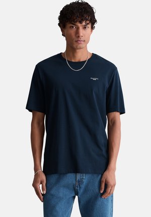 Junger Mann mit lockigem Haar trägt ein marineblaues T-Shirt mit kleinem Brustlogo und blaue Jeans, steht vor einem schlichten Hintergrund.