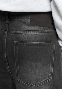 Los jeans de mezclilla gris presentan un bolsillo trasero, una etiqueta de cuero marrón con la marca y una textura suave con un ligero brillo.