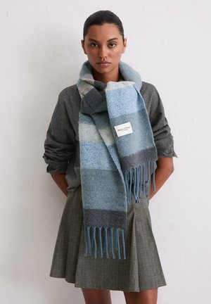 Blau-grau kariertes Wollschal mit Fransen, mit dem Label "Marc O'Polo", getragen über einem grauen Oberteil und einem olivgrünen Rock.