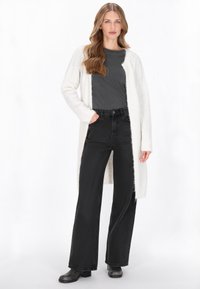 Hosszú, krémszínű kardigán puha textúrával, sötétszürke felső felett. Magas derekú, fekete, széles szárú farmernadrág és fekete blokk-sarkú csizmák.