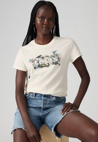 Tricou crem cu un logo floral Levi's pe față, asociat cu pantaloni scurți din denim bleu deschis, având margini destrămate.