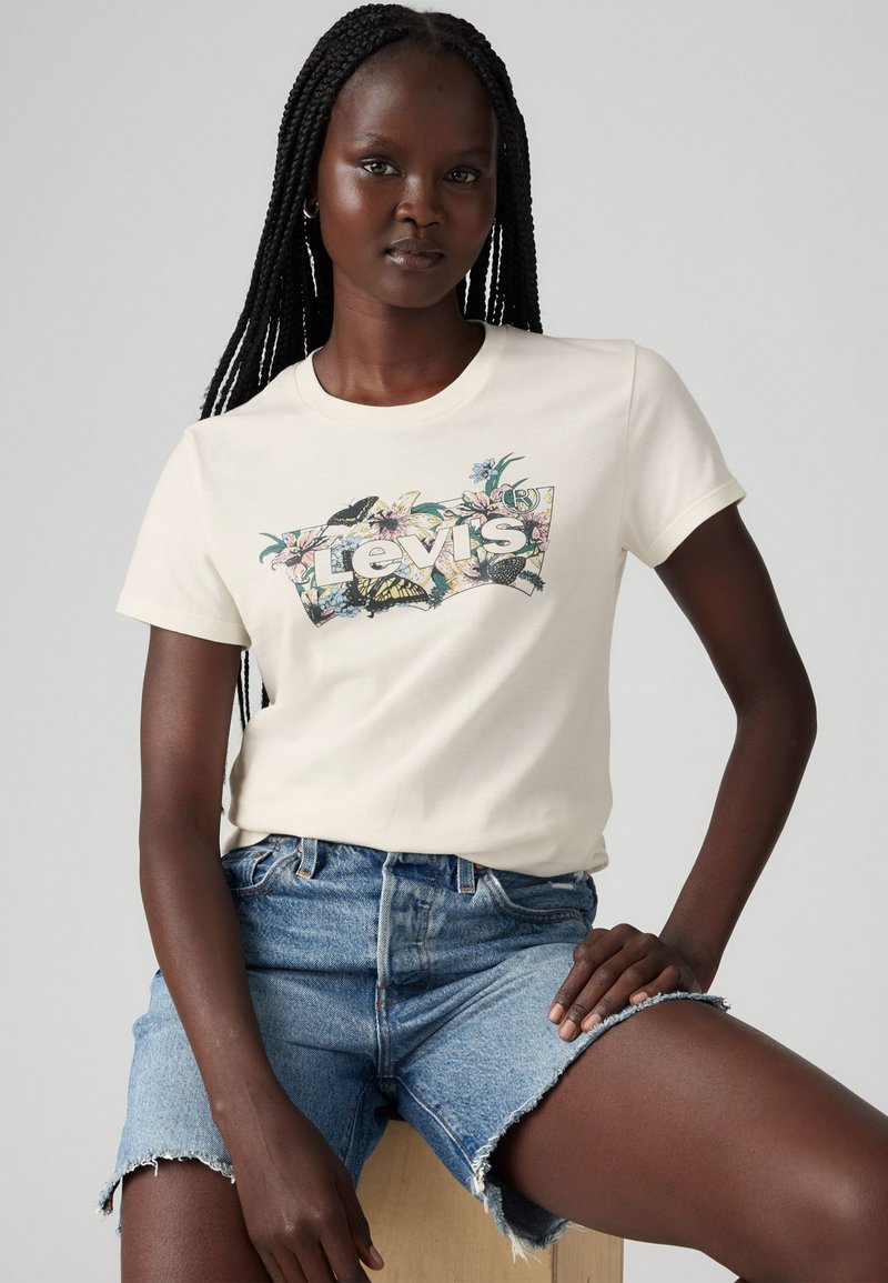 Tricou crem cu un logo floral Levi's pe față, asociat cu pantaloni scurți din denim bleu deschis, având margini destrămate.