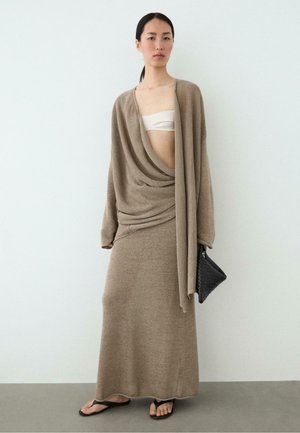 Frau trägt taupefarbenes, gestricktes, drapiertes Kleid mit langen Ärmeln, Bandeau-Oberteil, schwarze Flip-Flops und trägt eine schwarze, gewebte Handtasche, steht auf grauem Boden.