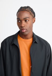 Sort lynlåsjakke med en blød mat finish over en lys orange crew neck skjorte. Modelens hår er flettet, med en neutral baggrund.