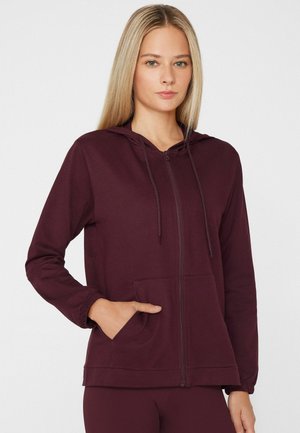 Frau mit langen blonden Haaren, die einen burgunderfarbenen Hoodie mit Reißverschluss und Kordeln trägt und eine Hand in die Fronttasche steckt.