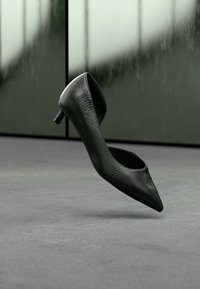 Μαύρο γοβάκι με μυτερή μύτη, που διαθέτει υφασμάτινο πάνω μέρος, χαμηλό τακούνι στιλέτο και ανοιχτή πλάτη, τοποθετημένο σε γκρι φόντο.