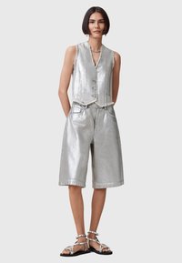 Gilet metallico argento con scollatura a V e cinque bottoni, abbinato a pantaloni corti metallici con tasche. Indossato con sandali argentati con cinturini.