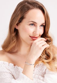 Zilveren armband met een gedraaid ontwerp gedragen om de pols, gecombineerd met een gestructureerde witte off-shoulder top en golvend haar.