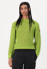 Suéter verde tricotado com uma malha solta, mangas compridas e gola redonda. Apresenta uma etiqueta vermelha na manga esquerda. Usado com calças pretas.