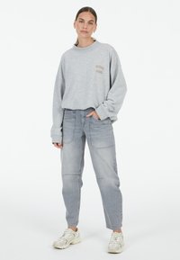 Gray oversized sweatshirt with a round neckline and embroidered "STUDIO AUREL" logo. Paired with light gray loose-fit jeans and beige sneakers.