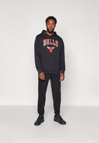 Felpa nera con "BULLS" e logo in rosso, abbinata a pantaloni della tuta neri e sneakers neri. Design semplice, materiale in cotone.