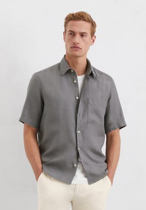 Short-sleeve, button-up shirt in grey linen with a single chest pocket, featuring a spread collar and subtle texture. Worn over a white tee.