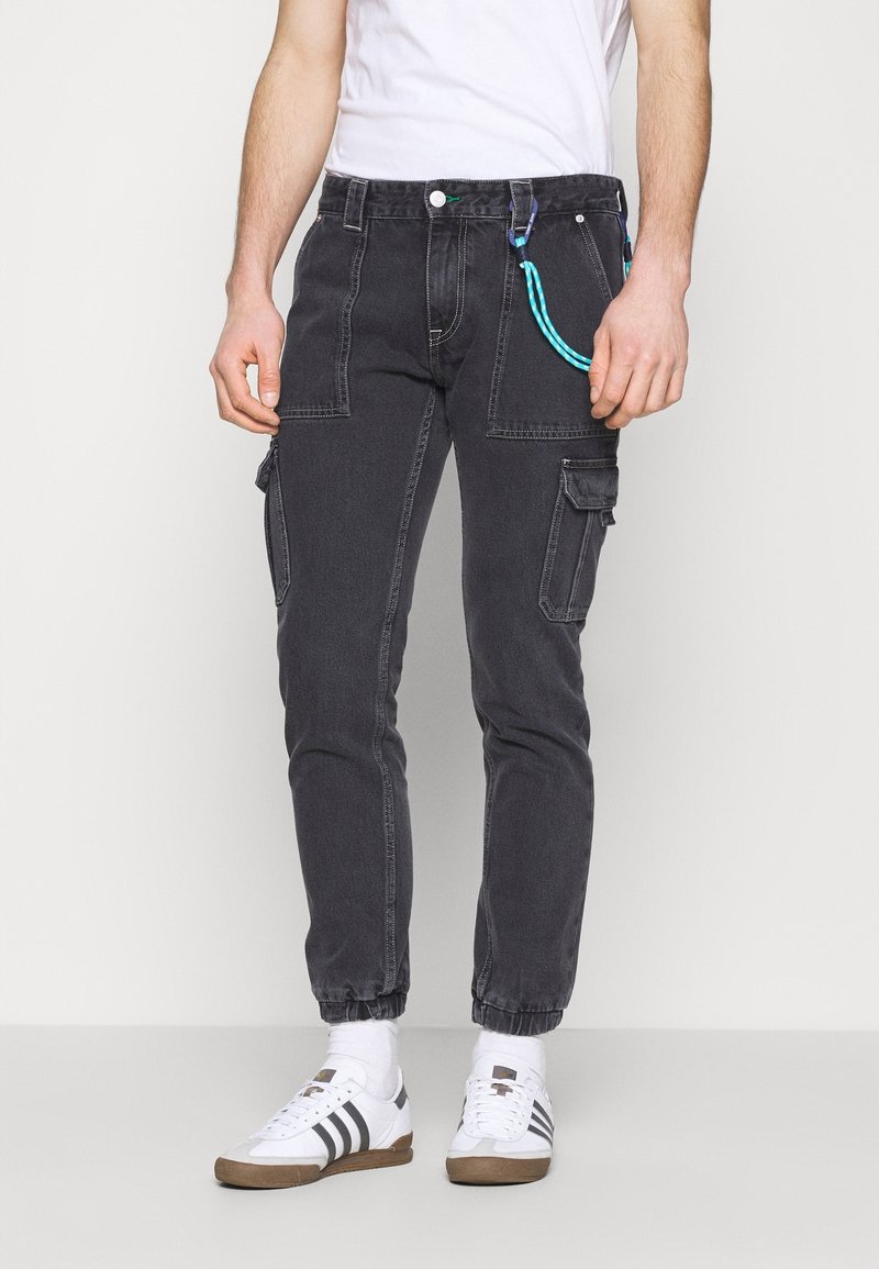Pantalones cargo en denim gris oscuro. Incluyen múltiples bolsillos y un lazo azul para accesorios. Pierna ajustada con puños elásticos. Combinados con zapatillas.