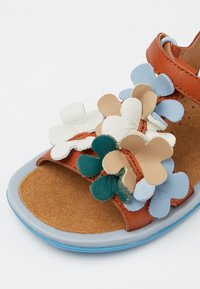 Sandalia de cuero marrón con aplicaciones de flores en capas en verde, azul, beige y blanco. Plantilla de ante suave y suela de goma azul.