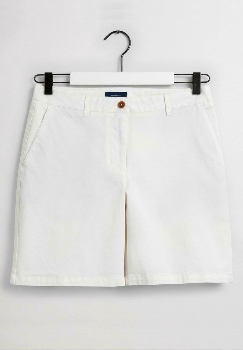 Vita knälånga shorts med framfickor, bälteshällor och en brun knapp, hängande på en svart och vit galge.