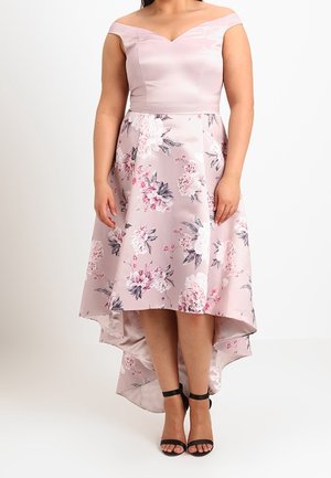 Vrouw draagt een roze off-shoulder jurk met een bloemenpatroon en een high-low zoom, gecombineerd met zwarte enkellaarsjes met enkelbandje, tegen een witte achtergrond.