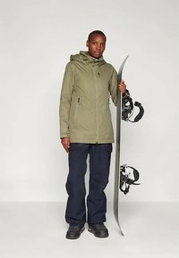 Casaco verde-azeitona com fecho éclair e capuz, combinado com calças de carga azul-marinho e botas de snowboard pretas, segurando um snowboard.