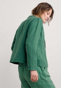 Vihreä pellavapuku, jossa on rento istuvuus, pyöreä kaula-aukko ja näkyvät tikkaukset. Yksinkertainen, koristamaton muotoilu, yhdistettynä yhteensopiviin housuihin.