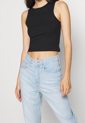 Female wearing sleeveless black crop top and light blue high-waisted jeans, standing with arms relaxed against a plain white background.