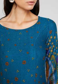 Femme portant un haut bleu à fleurs avec un col rond et une grande boucle d'oreille en perle, visible des épaules aux lèvres.