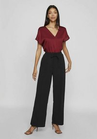 Mulher de pé, vestindo uma blusa bordeaux de decote em V, calças largas pretas de cintura alta e sandálias de salto castanhas, sobre um fundo cinza liso.