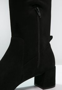 Black suede ankle boot with a block heel, side zipper, and smooth texture. Features a rounded toe and sleek design.