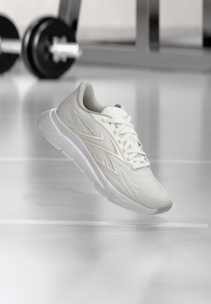 Zapatilla deportiva blanca con cordones flotando sobre un suelo reflectante de gimnasio, con una mancuerna y equipo desenfocados en el fondo.
