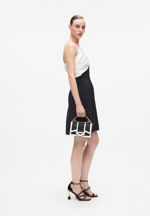 Zwart-witte avondjurk met een gedrapeerde witte top en een strakke zwarte rok. Model houdt een zwart-witte handtas met kettingaccenten vast. Zwarte strap-heeled schoenen.