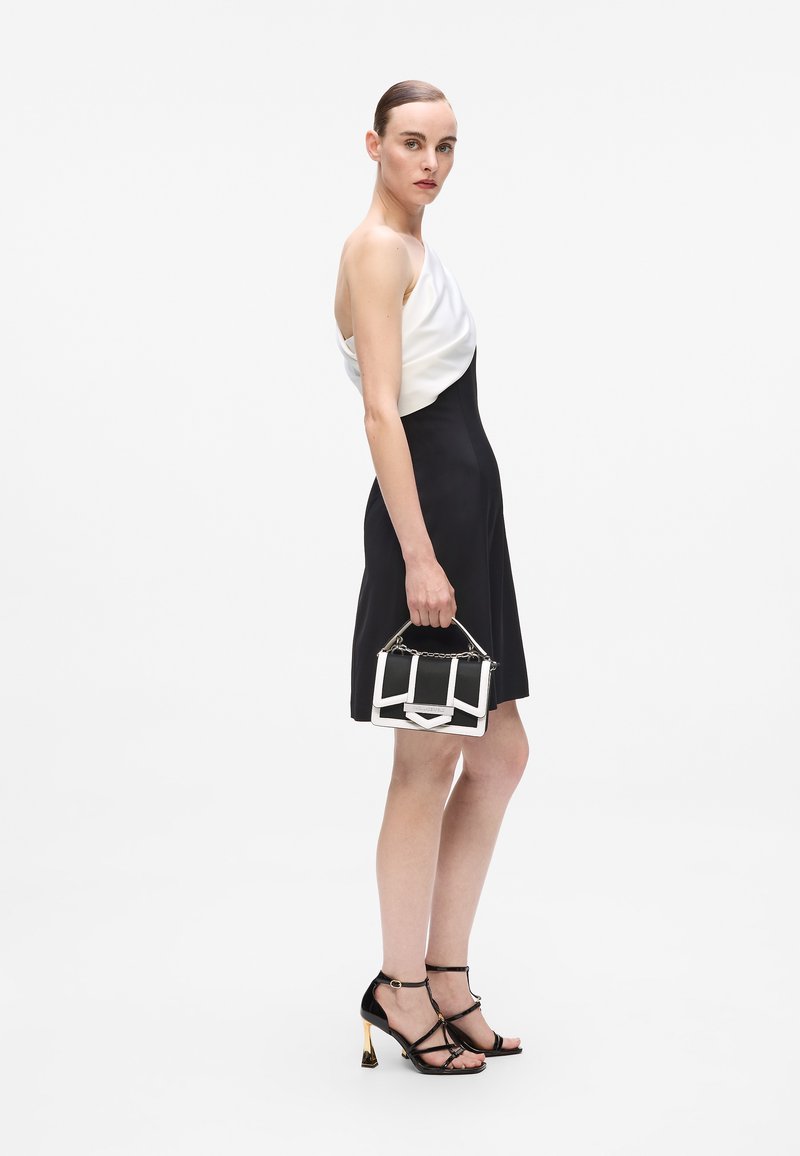 Zwart-witte avondjurk met een gedrapeerde witte top en een strakke zwarte rok. Model houdt een zwart-witte handtas met kettingaccenten vast. Zwarte strap-heeled schoenen.
