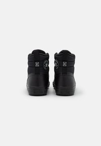 Black mid-top sneakers with textured fabric and rubber material. Features logo patches at the heel and subtle stitching details.