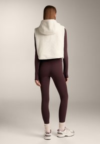 Gekookte wol vest in off-white met een capuchon, gecombineerd met strakke donkerburgundy leggings en lichte sportieve schoenen met paarse accenten.