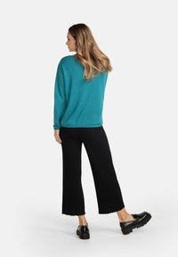 Türkiser Pullover mit langen Ärmeln, strukturierter Stoff, kombiniert mit schwarzen, gerippten, weit geschnittenen Hosen und klobigen schwarzen Schuhen. Schlichter Hintergrund.